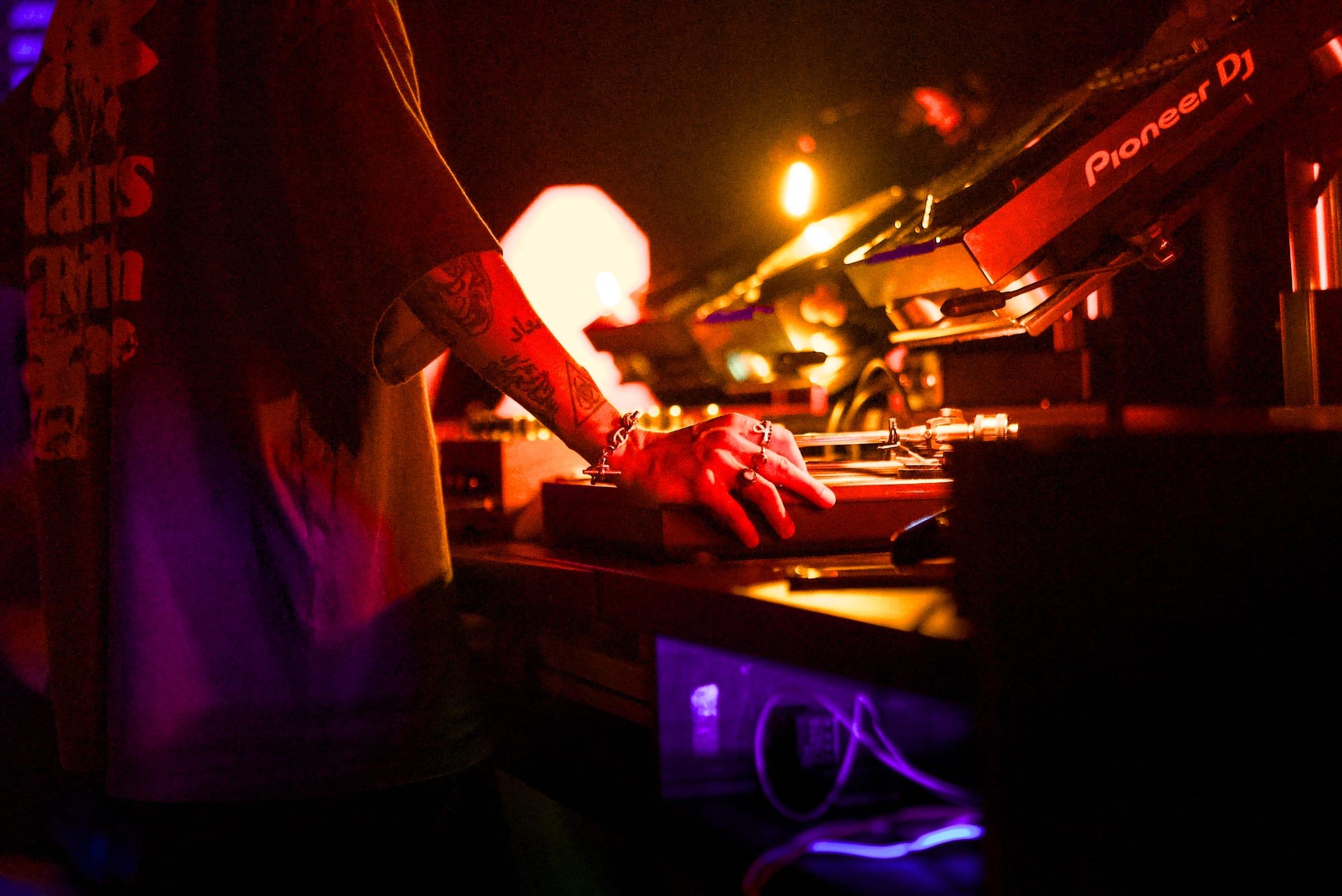 Close-up of a tattooed hand resting on a Pioneer turntable, lit in warm amber light.