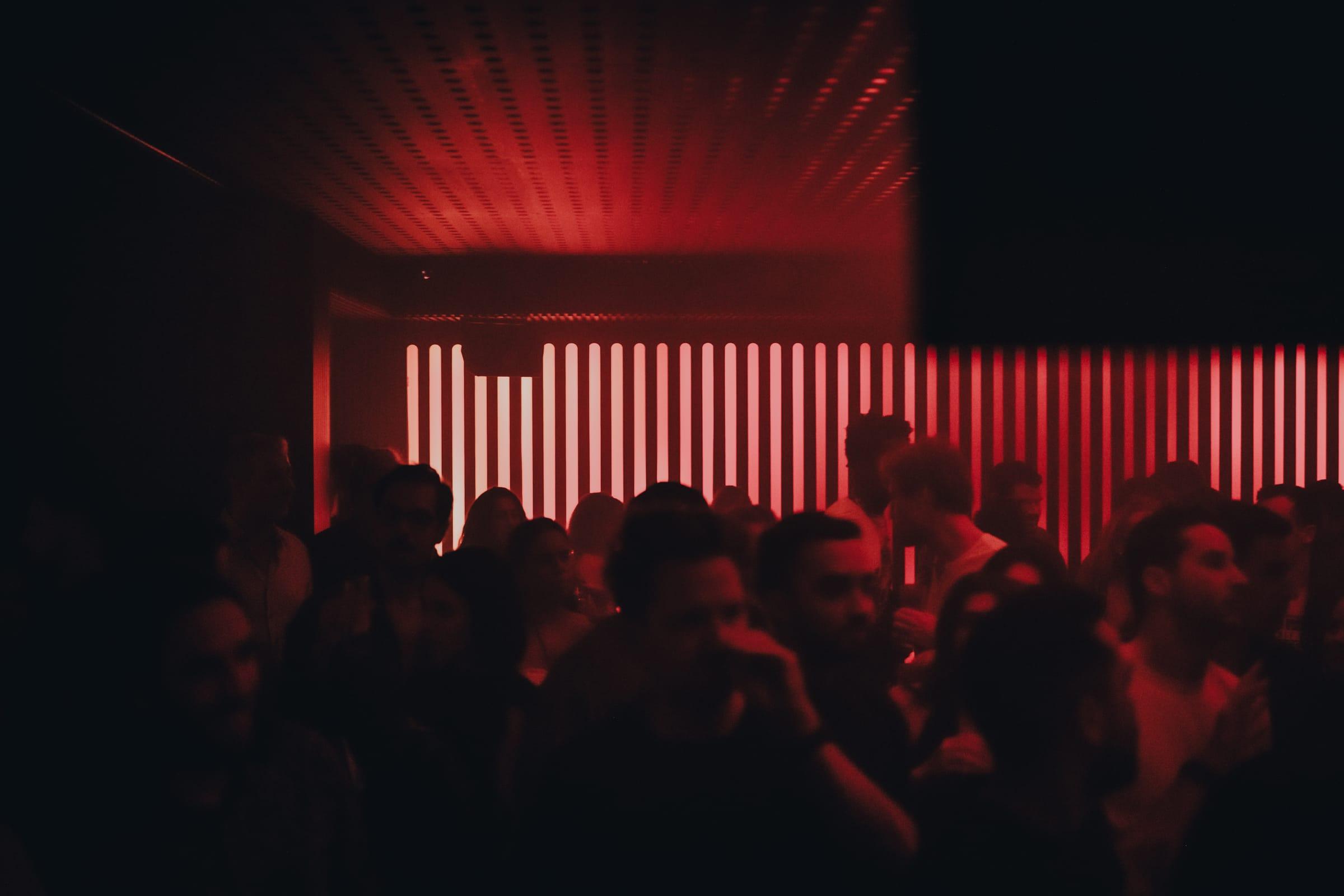 A crowd dances with hands raised beneath the red dot-ceiling, the DJ booth glowing behind them.