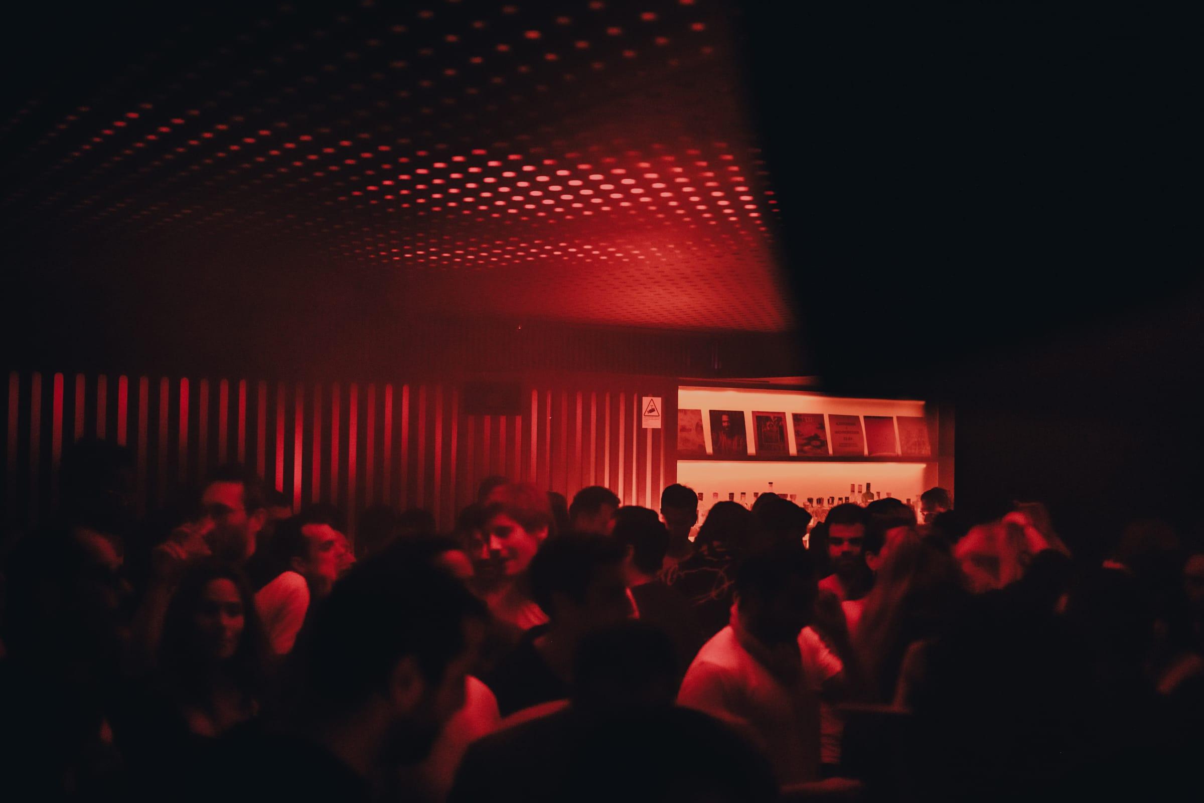 A crowd gathers beneath the red dot-perforated ceiling, a wall of framed record sleeves glowing warm behind them.