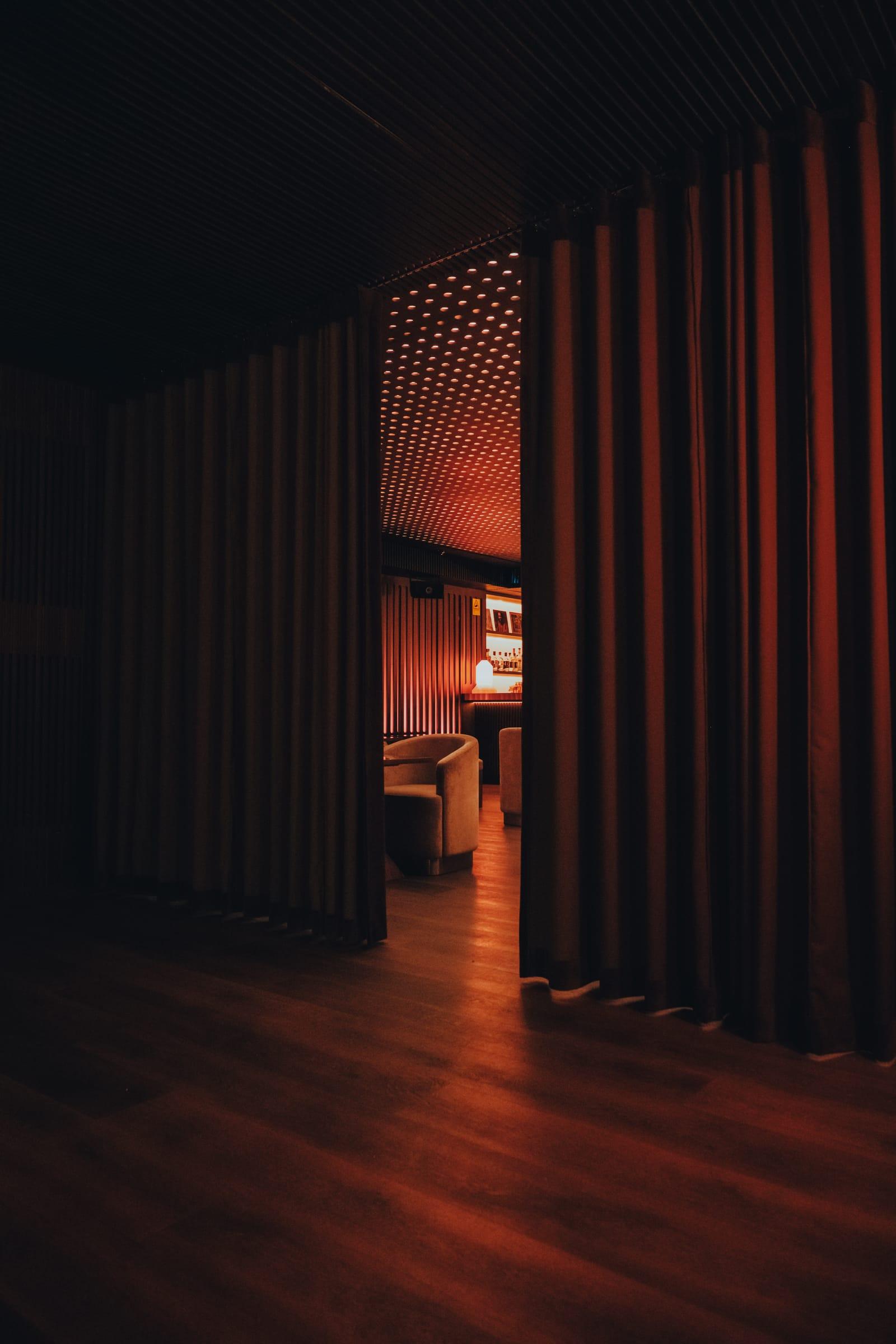A heavy curtain parts onto a warmly lit room beyond, its perforated ceiling glowing red above a lounge chair and the back bar.