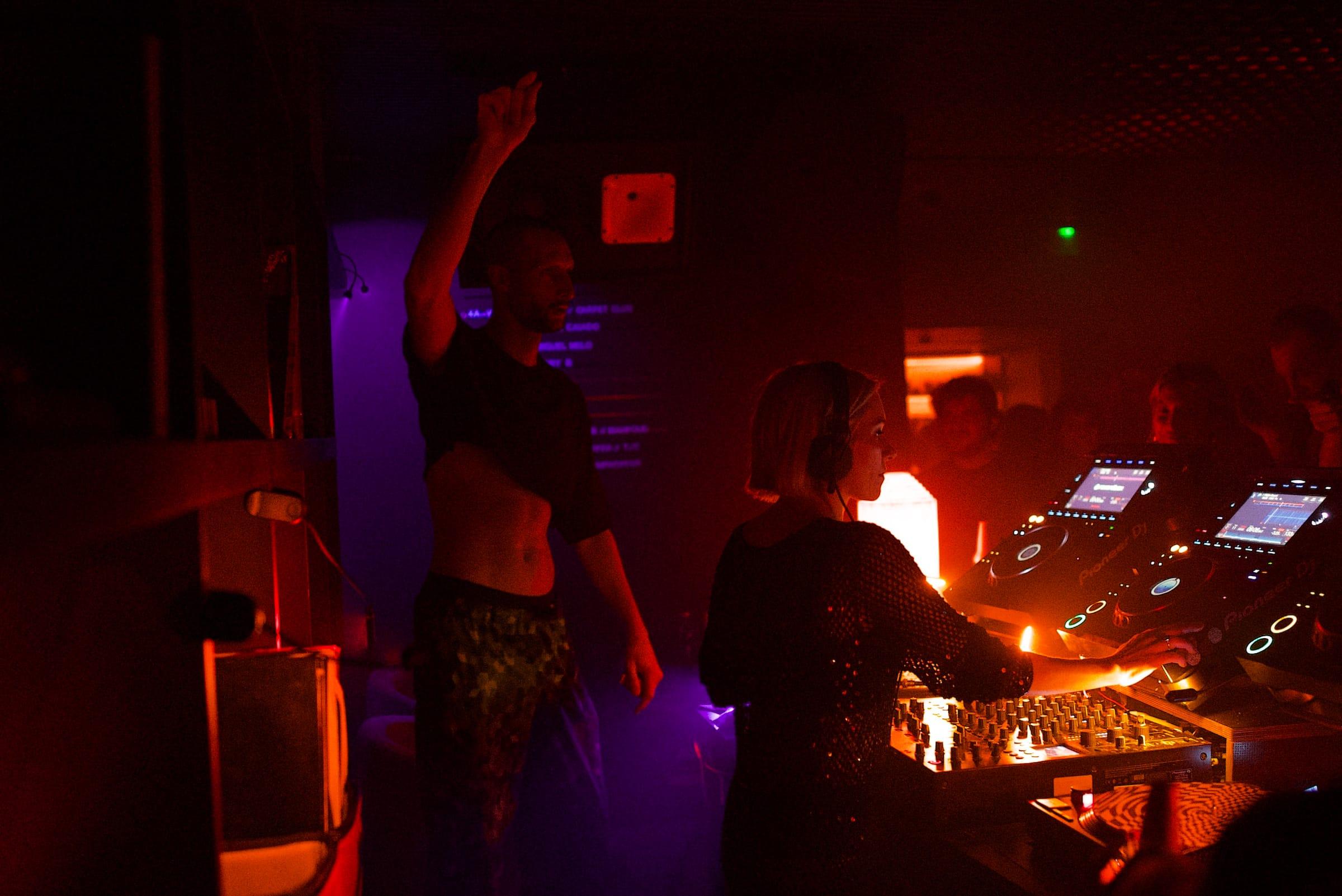 Two people at the DJ booth, one with an arm raised mid-dance, purple stage light glowing behind them.