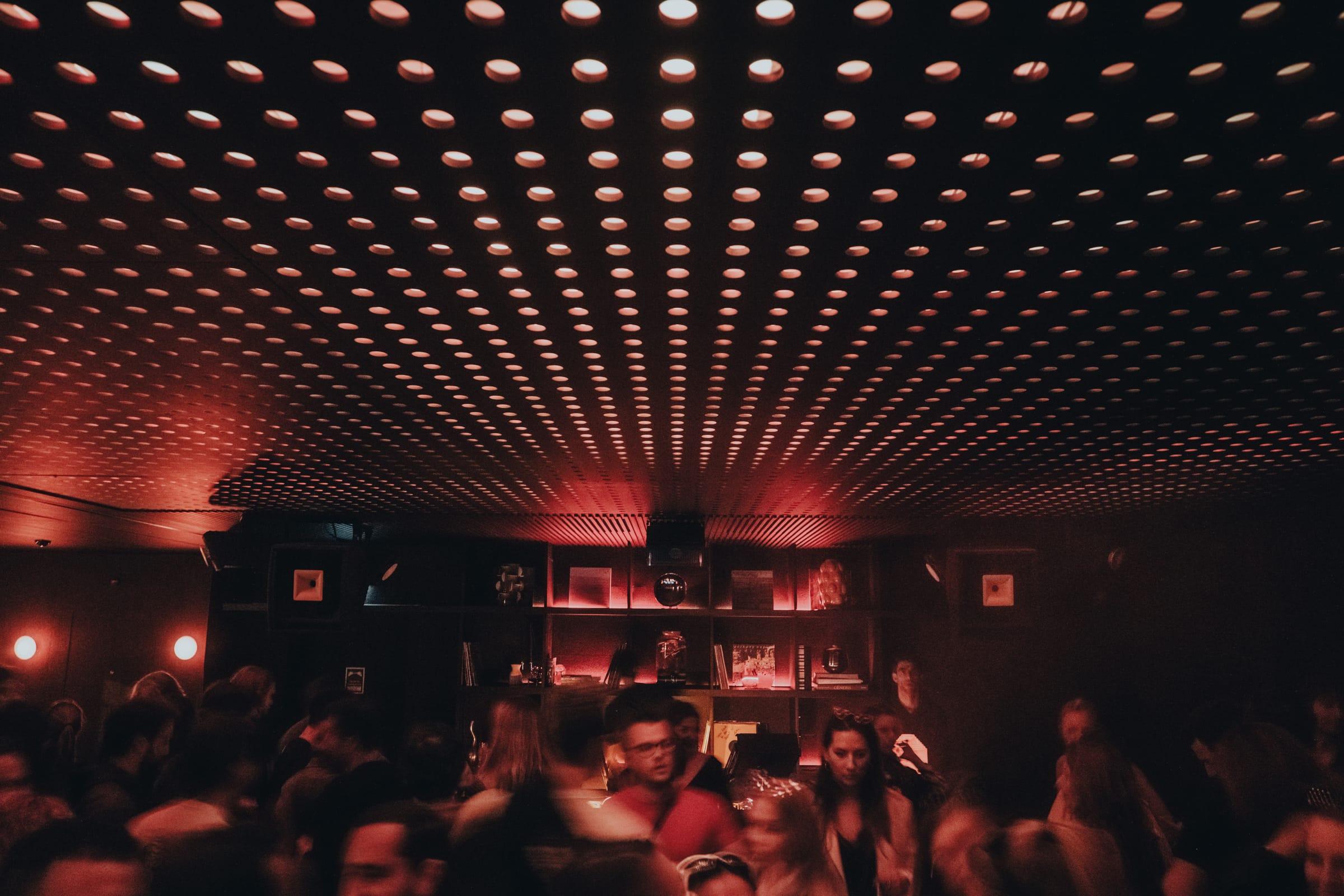 A DJ mixes at the decks under the red dot-ceiling, flanked by a shelf of records and sculptural objects.