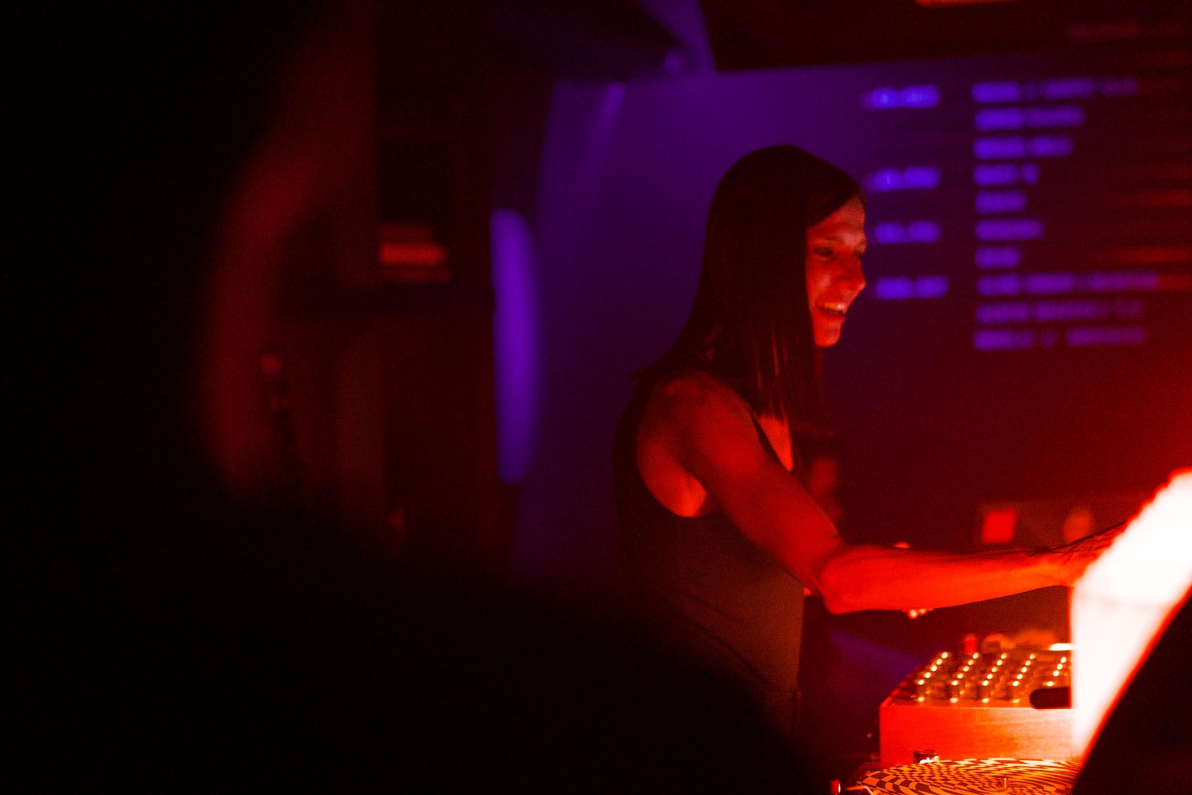 A DJ smiles while mixing, lit in purple and orange stage light.