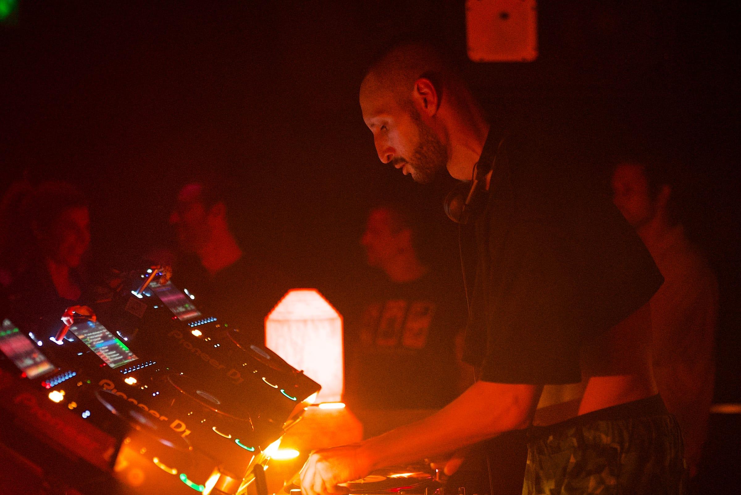 A DJ leans over the decks, headphones around his neck, the crowd softly lit in warm orange behind him.