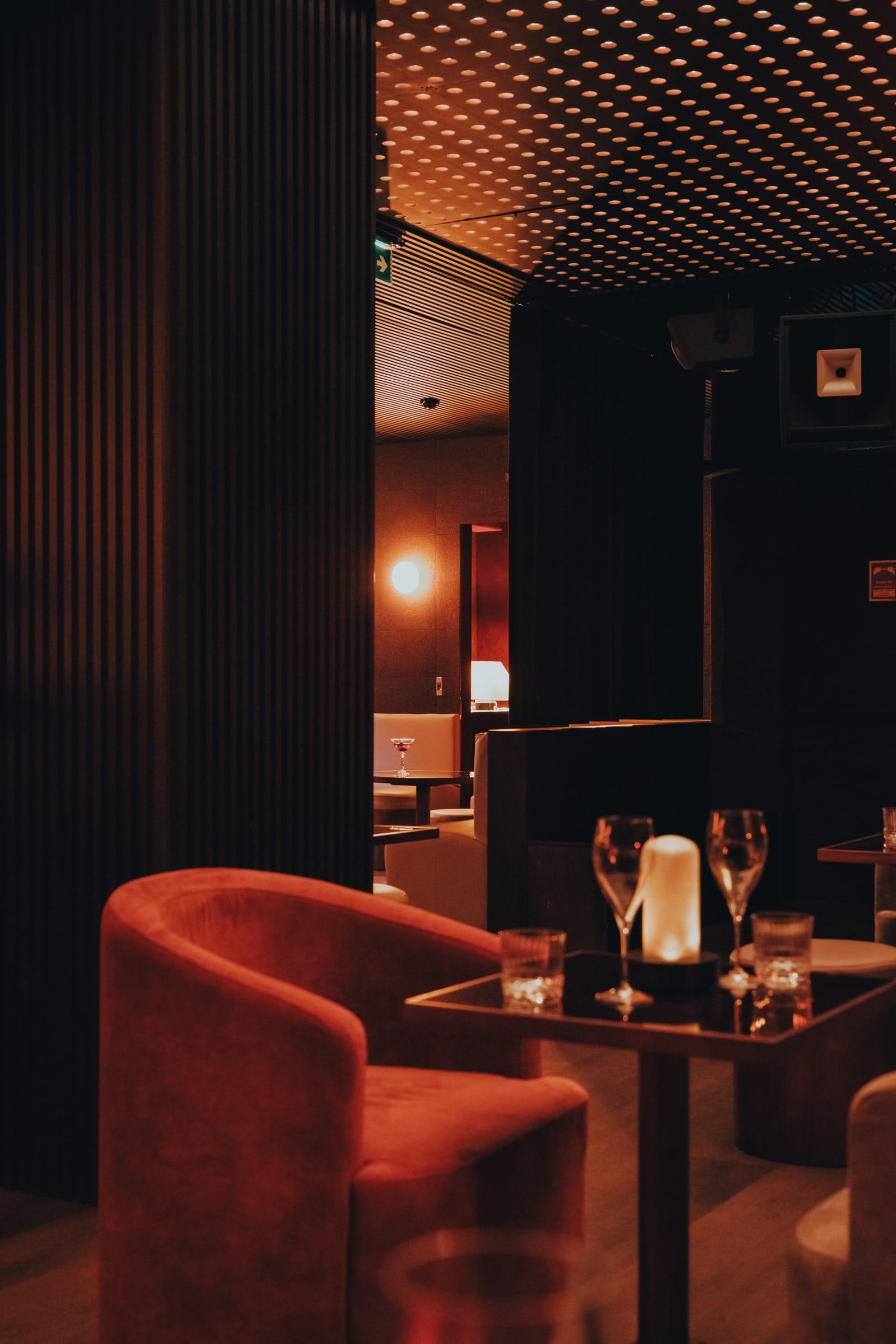 A curtain frames a doorway into a red dot-lit lounge, a rose velvet armchair and a candlelit table with wine glasses in the foreground.