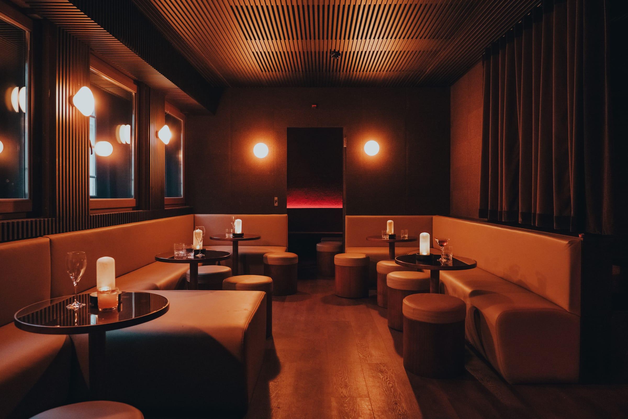 A curved lounge banquette lined with round tables, lit candles, and cocktail glasses, framed by warm round wall sconces.