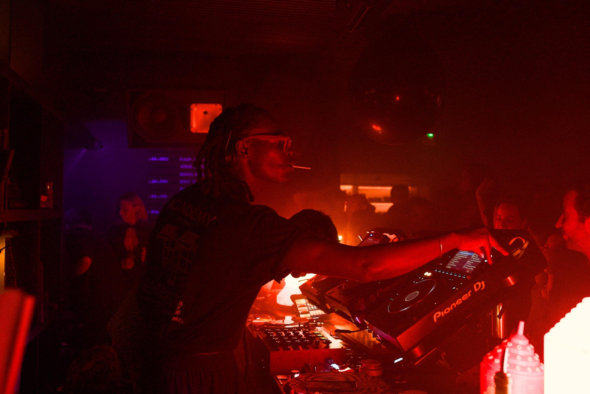 A DJ in red-tinted glasses works the decks with a cigarette in his mouth, a disco ball turning above him in thick red haze.