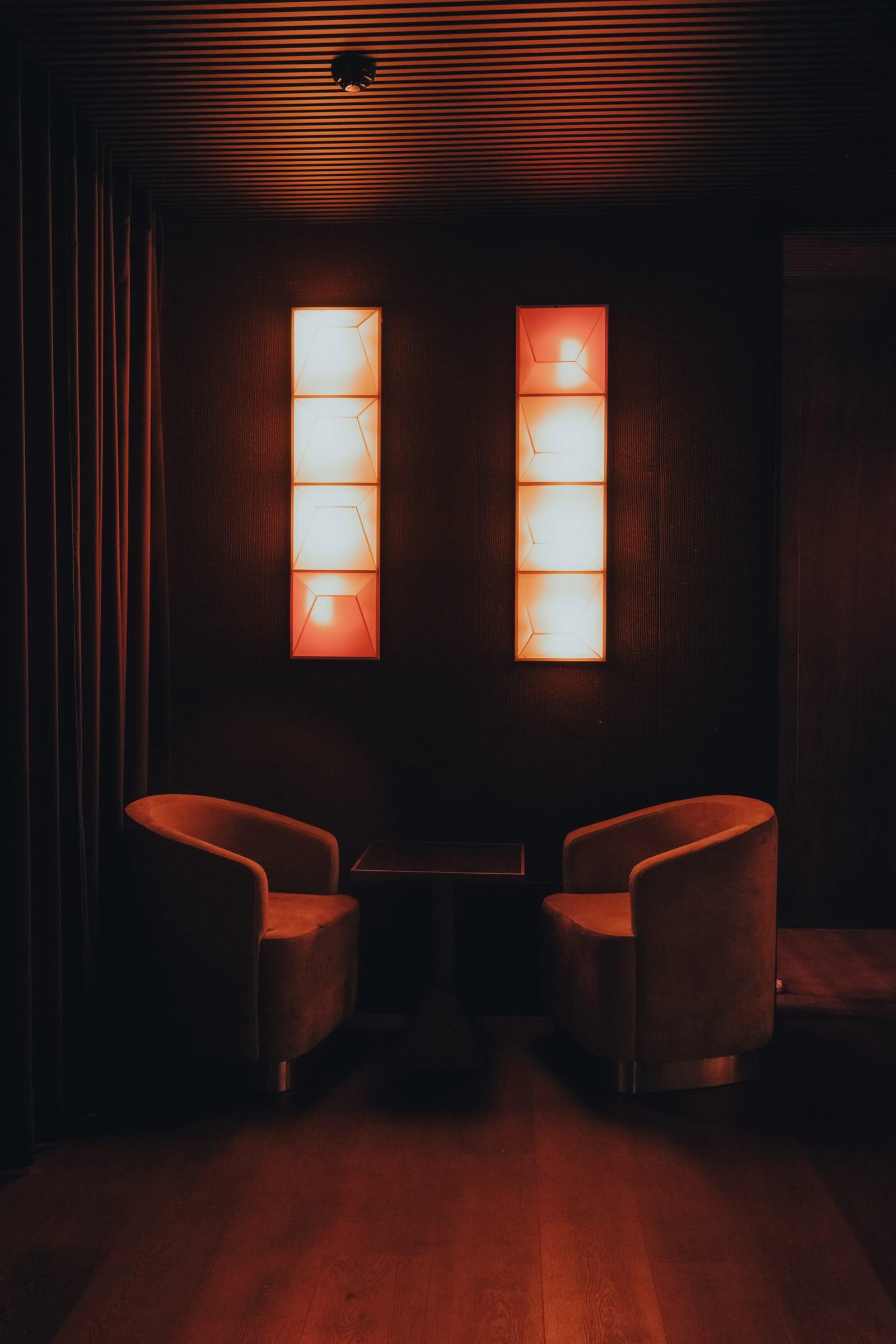 Two velvet armchairs face each other around a small table in a dim red-lit corner, lit by a pair of tall glowing wall panels.