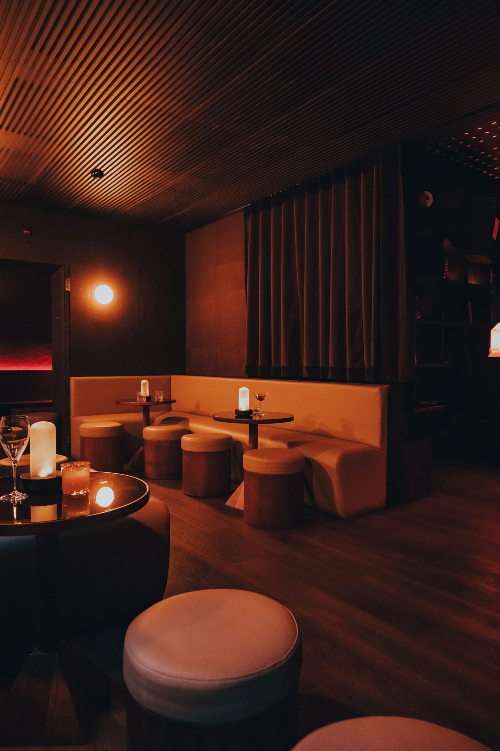 A curtain parts onto a warmly lit lounge beyond, round candlelit tables with wine glasses beside a cream banquette under a wood-slat ceiling.